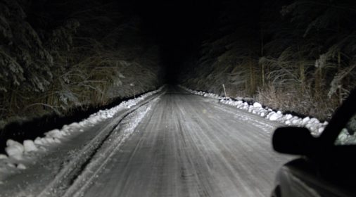 dark road with snow on it with trees either side, taken out of car window