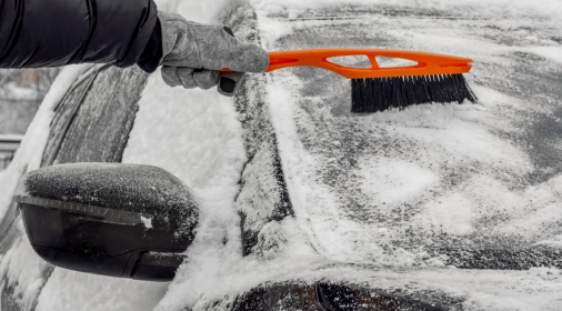 Man using Car Snow Brush to remove snow from the car windshield. Person in a black jacket and gray gloves. Close-up. Trees are covered with snow in the background. Winter, daytime.