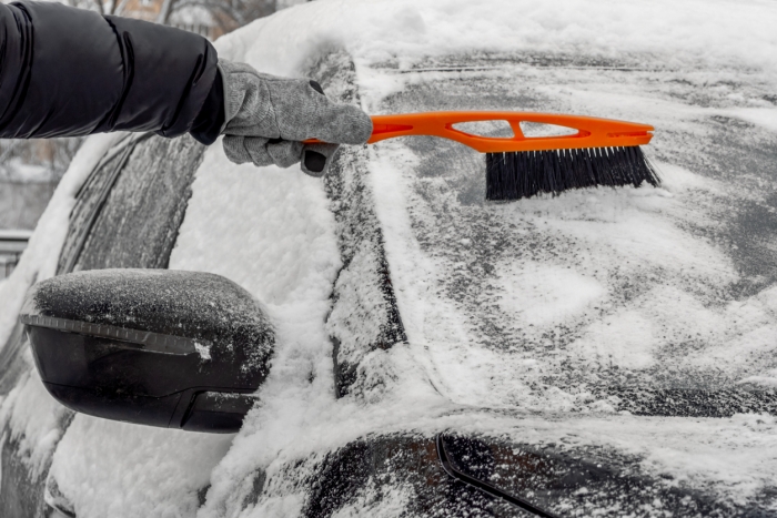 Man using Car Snow Brush to remove snow from the car windshield. Person in a black jacket and gray gloves. Close-up. Trees are covered with snow in the background. Winter, daytime.