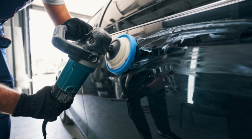 Worker polishing black car side with polisher while holding instrument with both hands in repair service workshop